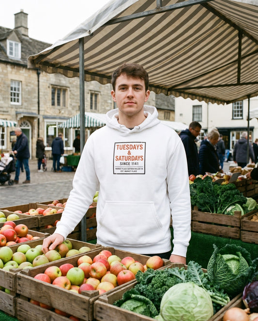 SINCE 1141 hoodie in white, worn behind a Saturday market stall of apples and winter greens – WALLED IN Saffron Walden. AI-styled photo. Real product, printed to order.
