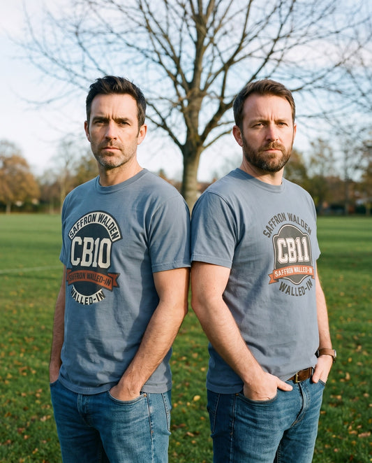 POSTCODE PRIDE CB11 and CB10 tees in blue jean, two friends back-to-back on a grassy common – WALLED IN Saffron Walden. AI-styled photo. Real product, printed to order.