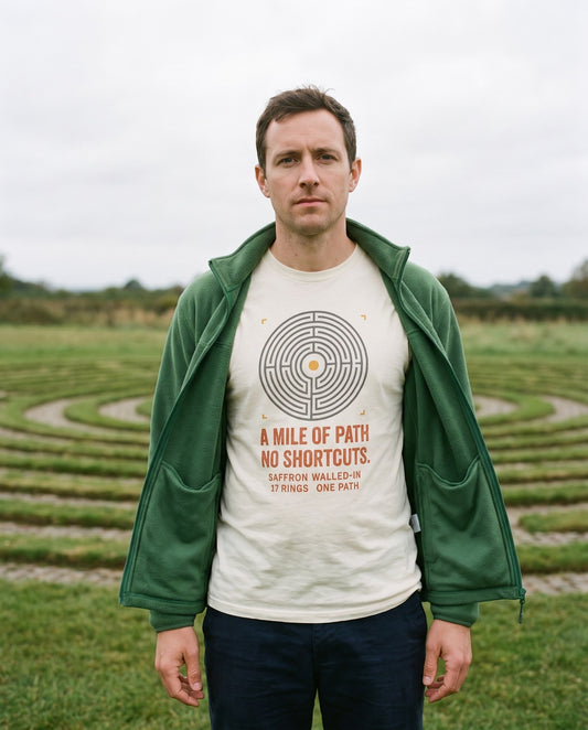 NO SHORTCUTS turf maze tee in ivory, worn with a green fleece beside a turf maze on a common – WALLED IN Saffron Walden. AI-styled photo. Real product, printed to order.