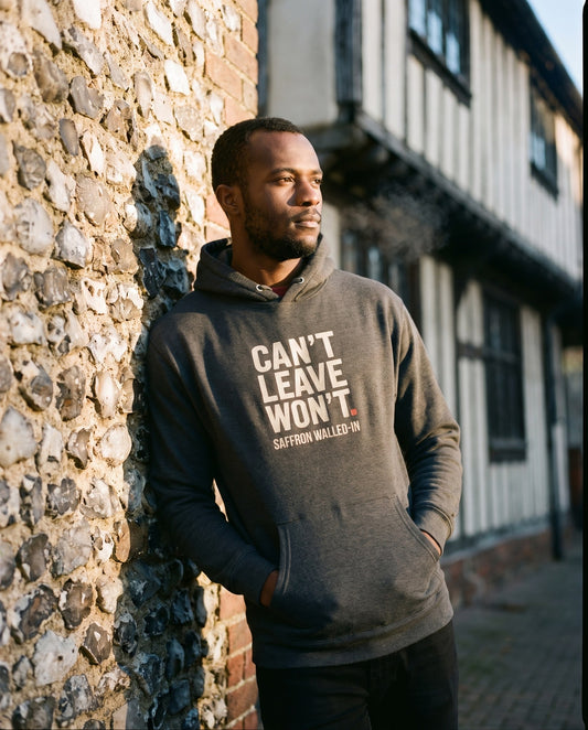CAN'T LEAVE. WON'T. hoodie in charcoal heather, worn by a man leaning on a flint wall in a market town – WALLED IN Saffron Walden. AI-styled photo. Real product, printed to order.