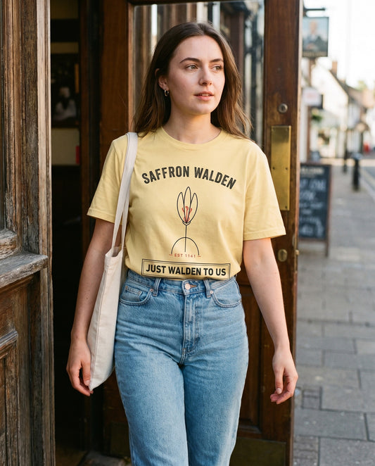 JUST WALDEN tee in butter, tucked into jeans, walking out of a timber-framed pub doorway – WALLED IN Saffron Walden. AI-styled photo. Real product, printed to order.